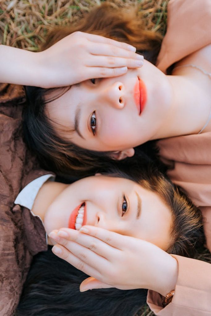 Two women with red lips and natural makeup smiling while lying on grass.