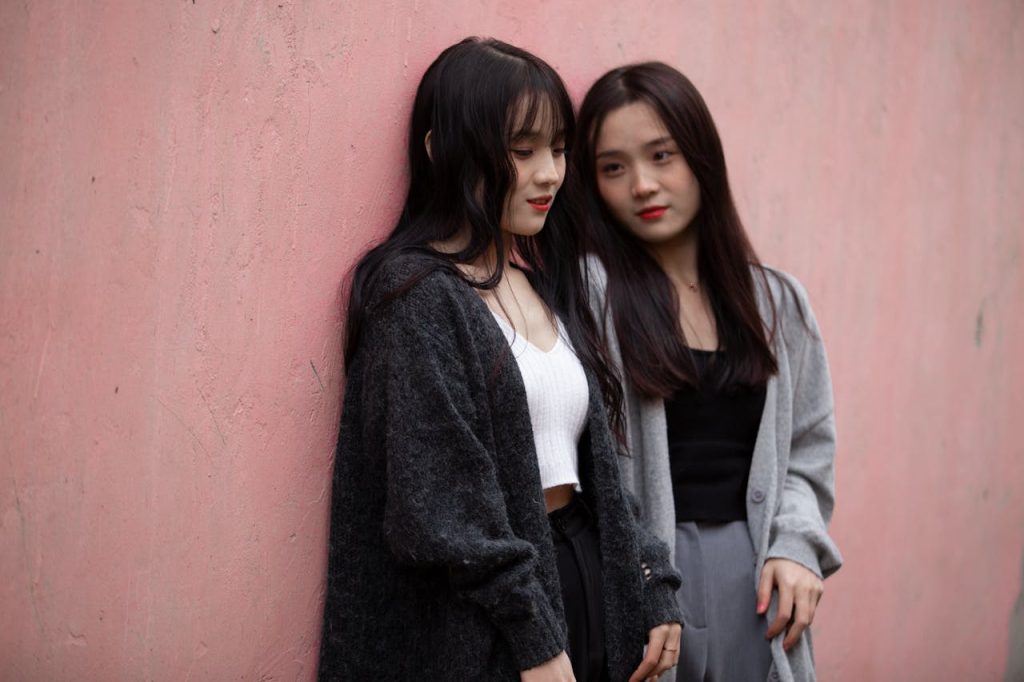 Two young women stand casually against a pink wall, sharing a moment of friendship and style.